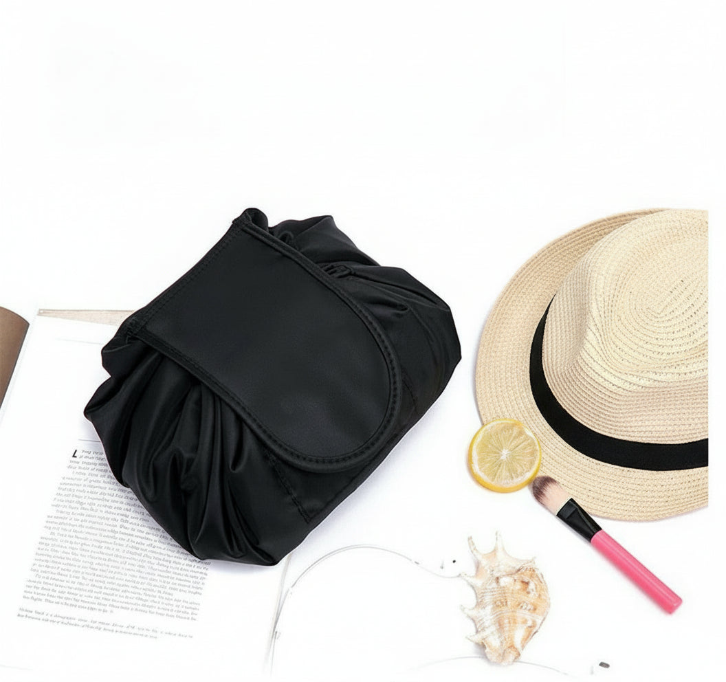 Black ruched handbag, straw hat with black band, lemon slice, shell, and pink makeup brush on a white surface.